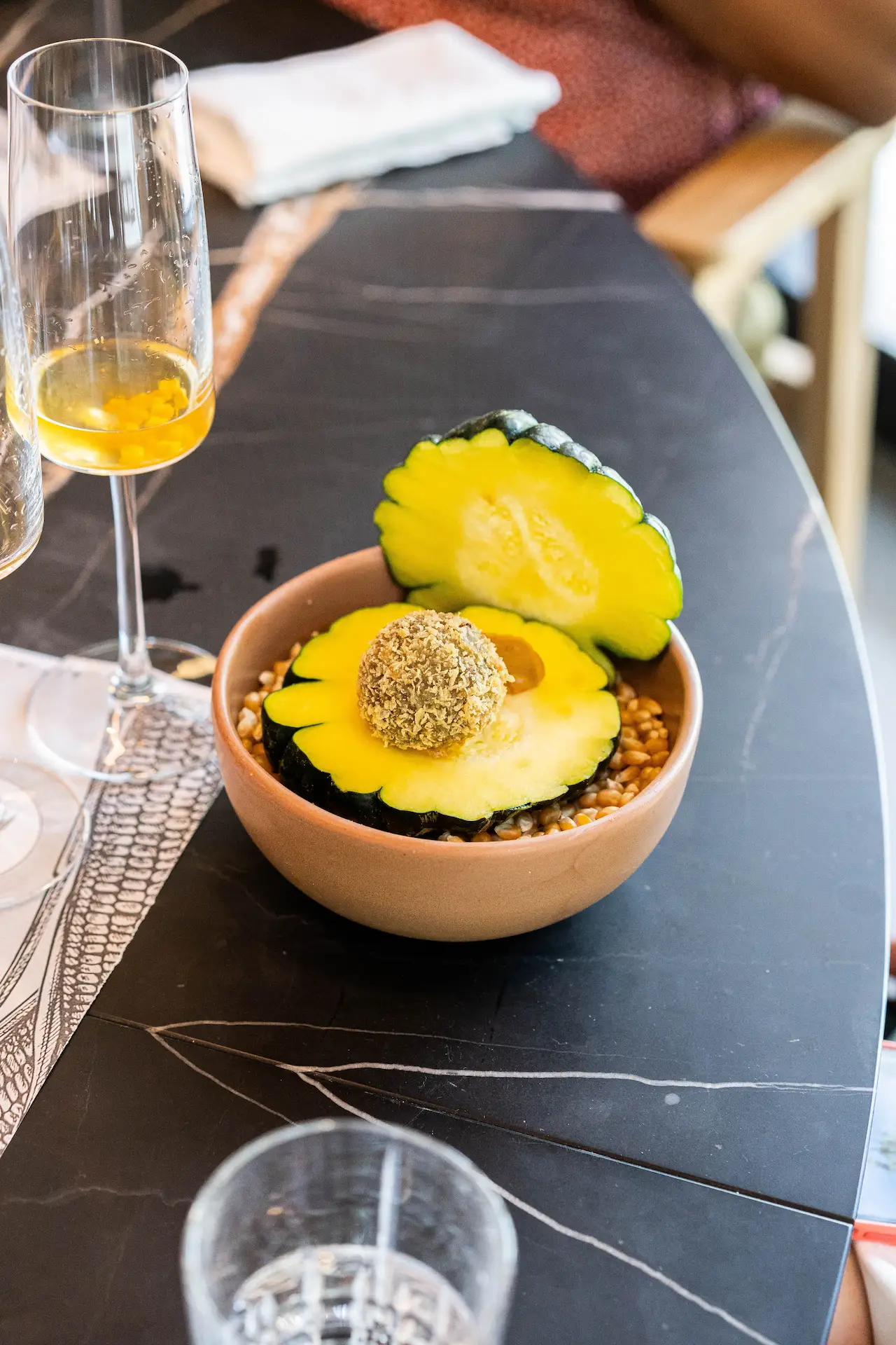 Squash and croquette dish served over grains in ceramic bowl with drinks on marble table