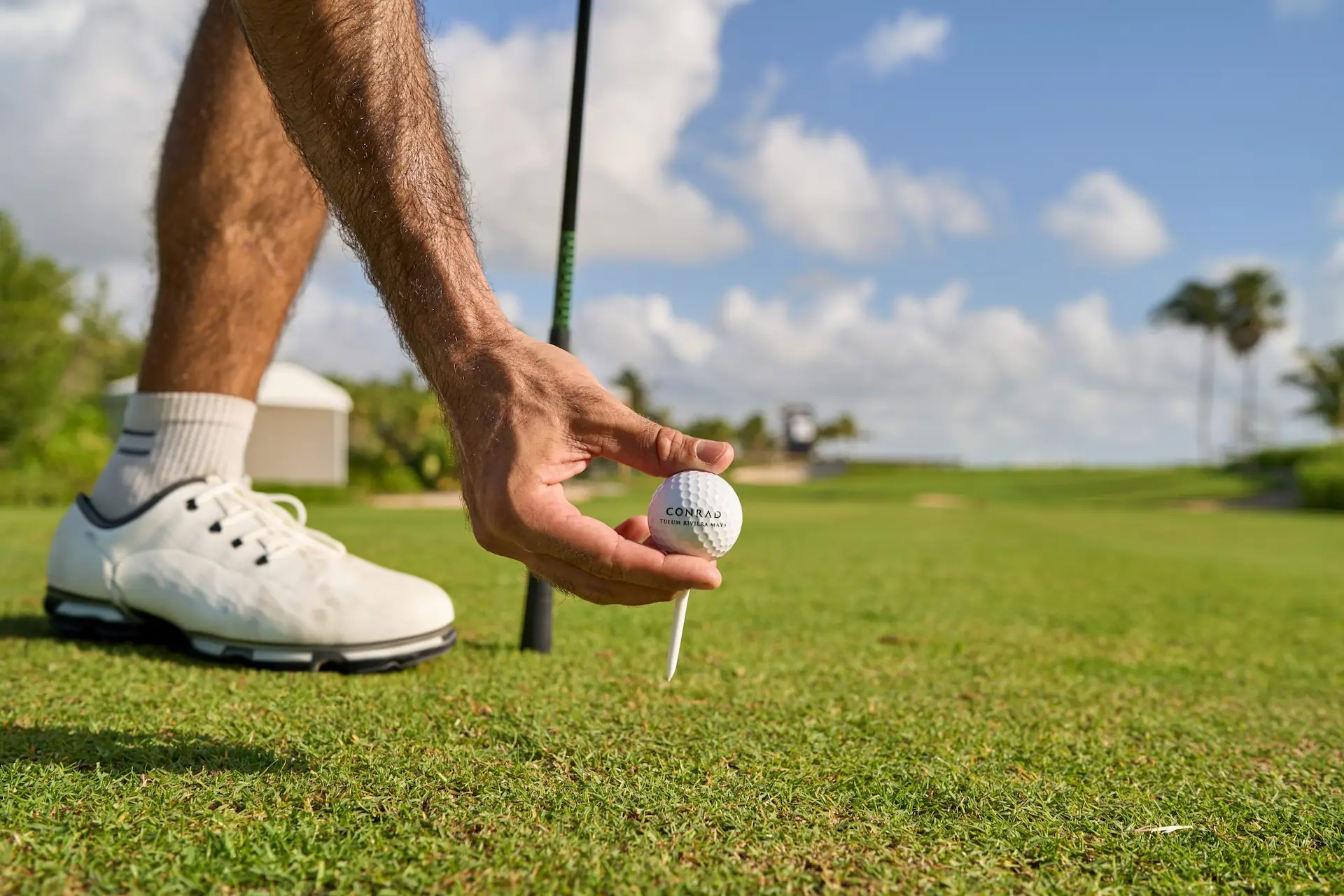 Golf ball on a tee held by a person in white golf shoes on a sunny course with palm trees.