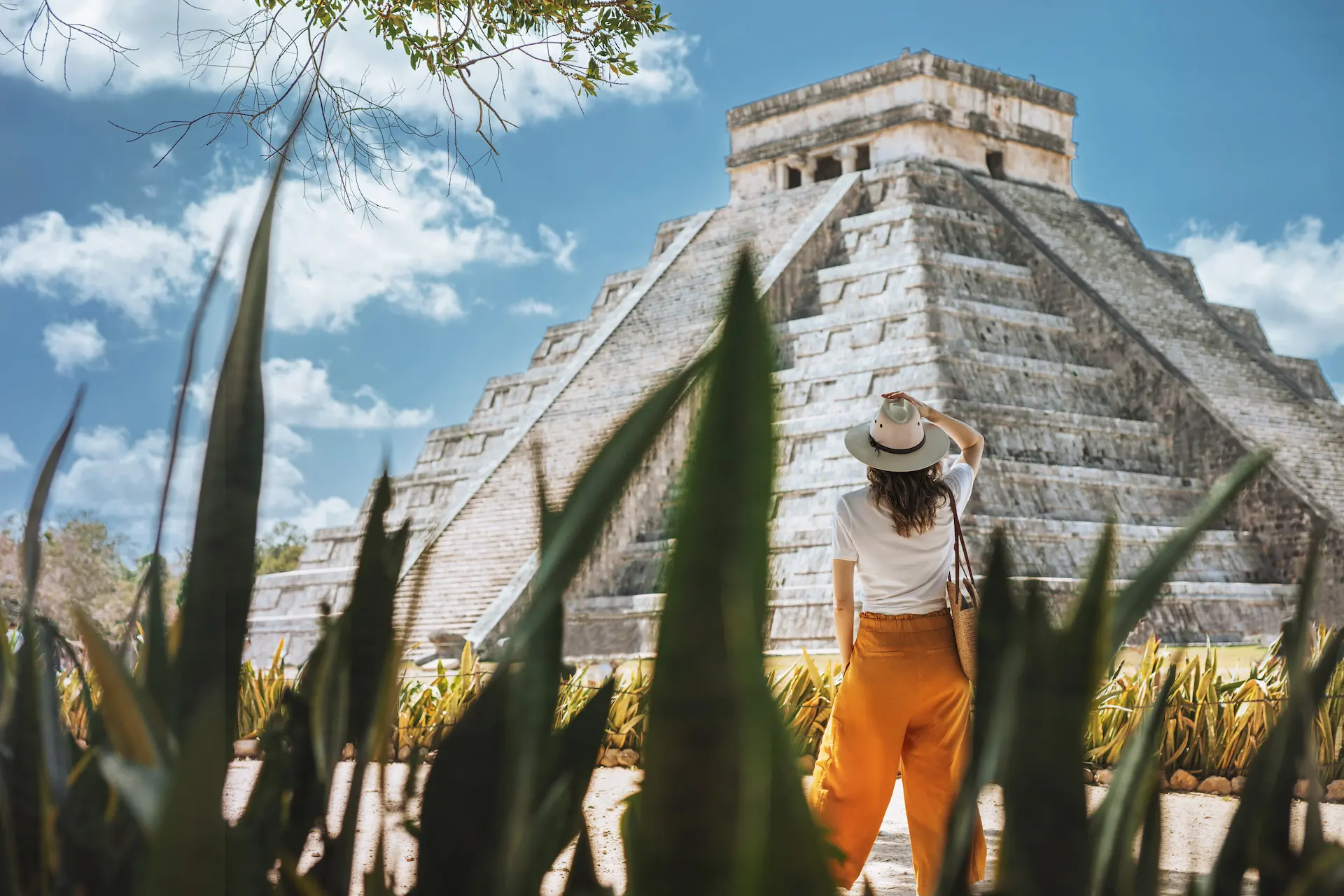 Person in white shirt and orange pants stands before Chichen Itza pyramid, framed by plants and blue sky.