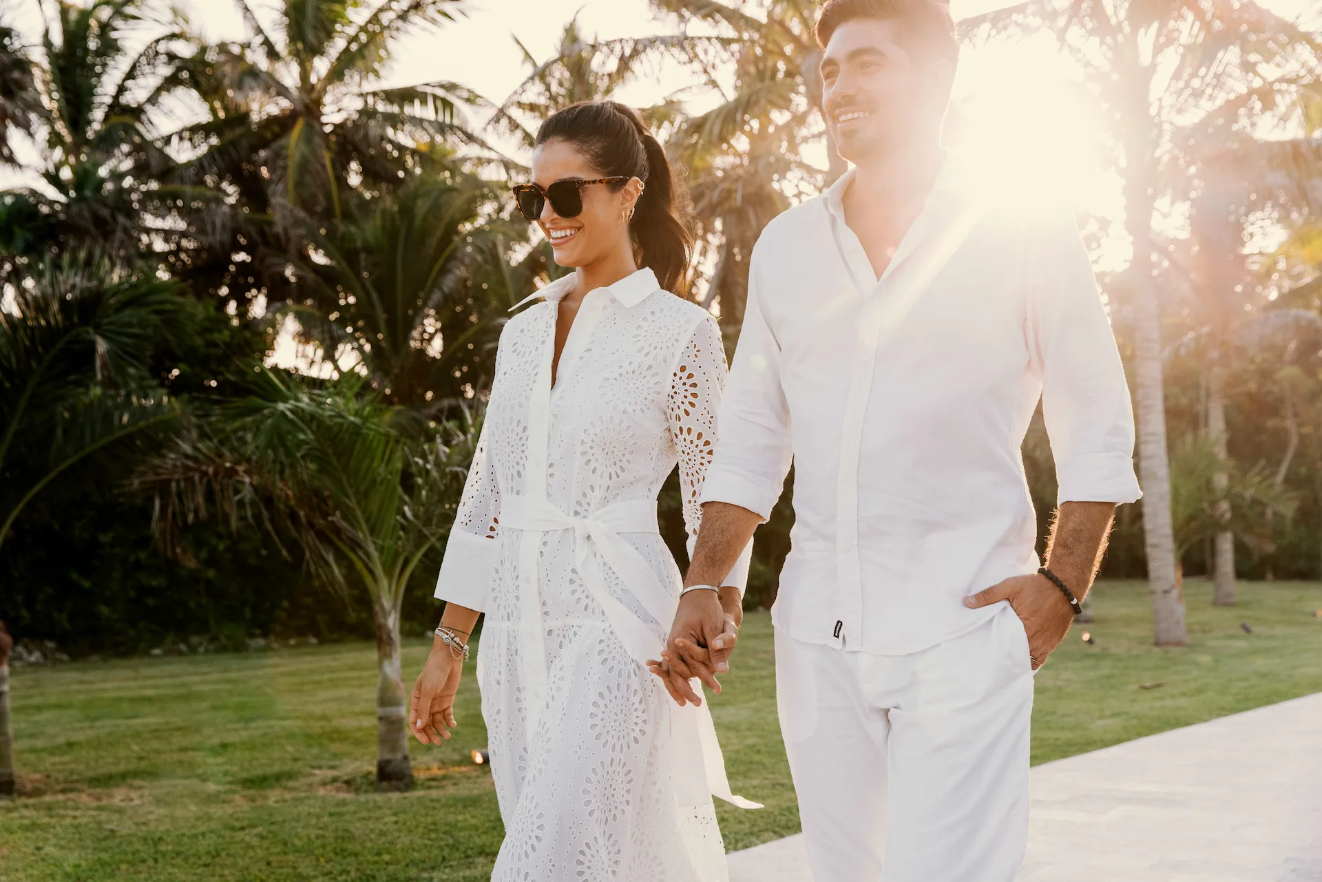 Two people in white walk hand-in-hand through a tropical garden, sunlight filtering through palm trees.