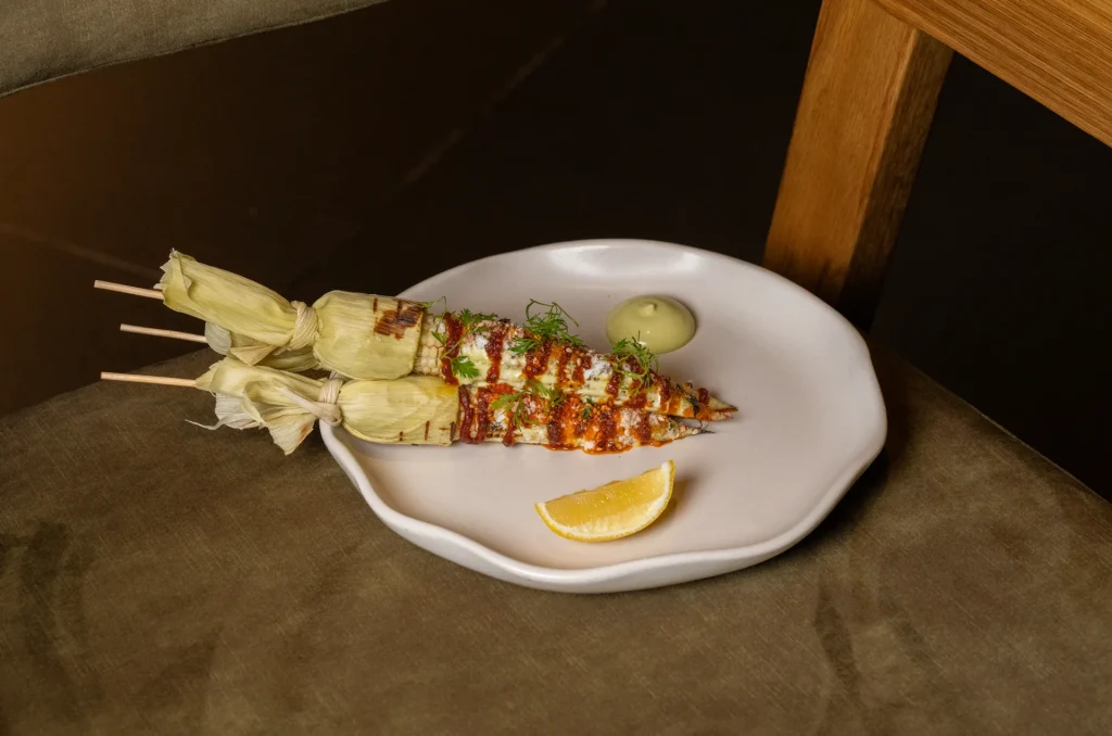 Grilled corn on skewers with creamy sauce, herbs, cheese, green dip, and lemon wedge on plate