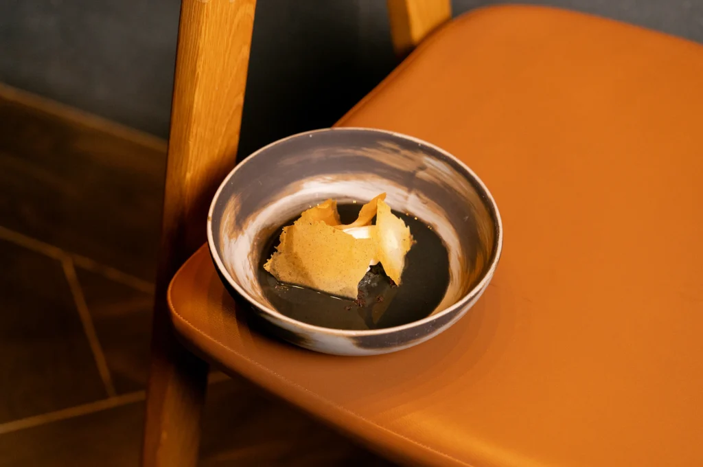 Gourmet dish with dark sauce, crispy golden garnish, and white cream in ceramic bowl on tan chair