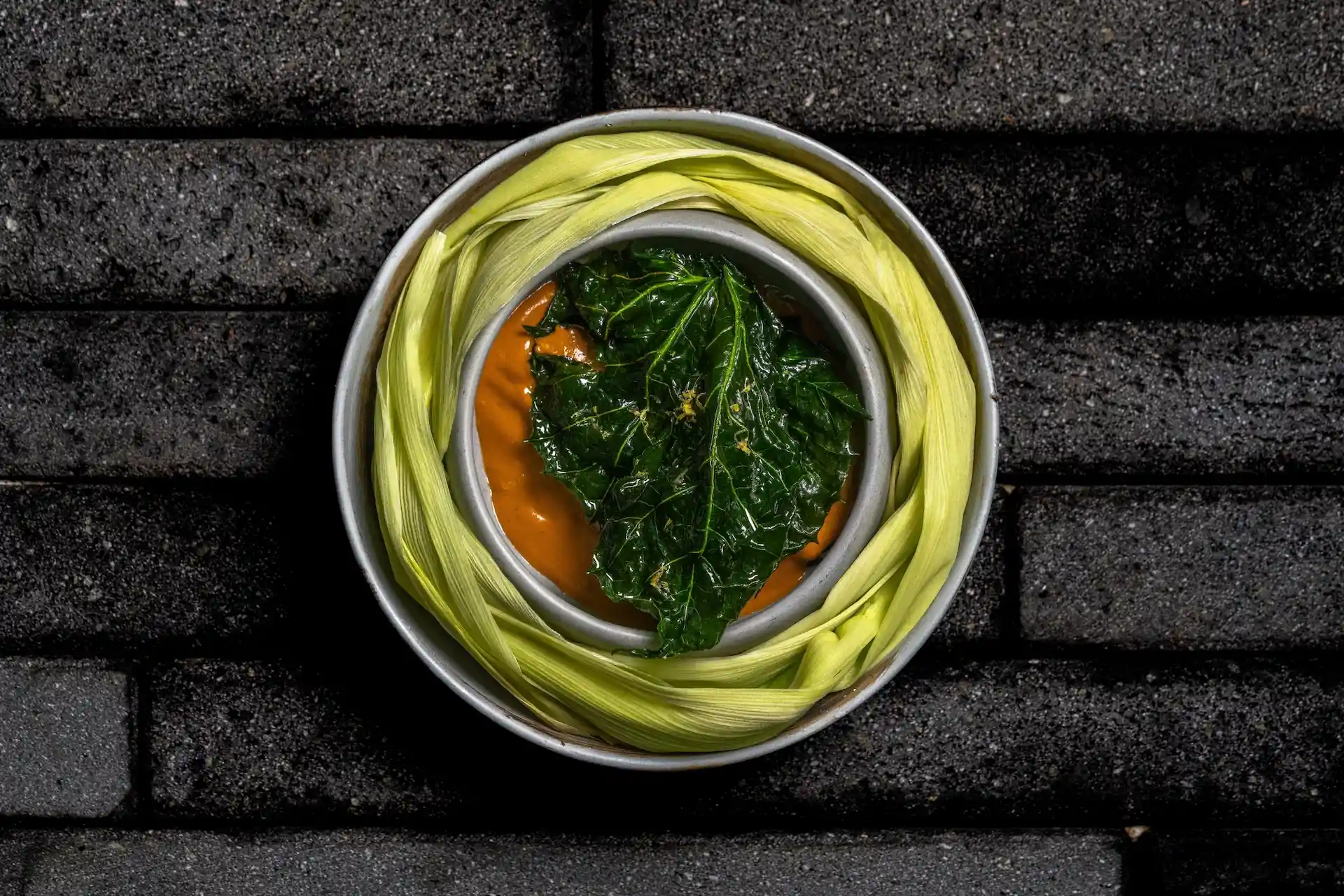 Plato con salsa de color naranja brillante cubierto con una hoja verde en un tazón rodeado de hojas de maíz dispuestas sobre una superficie oscura.