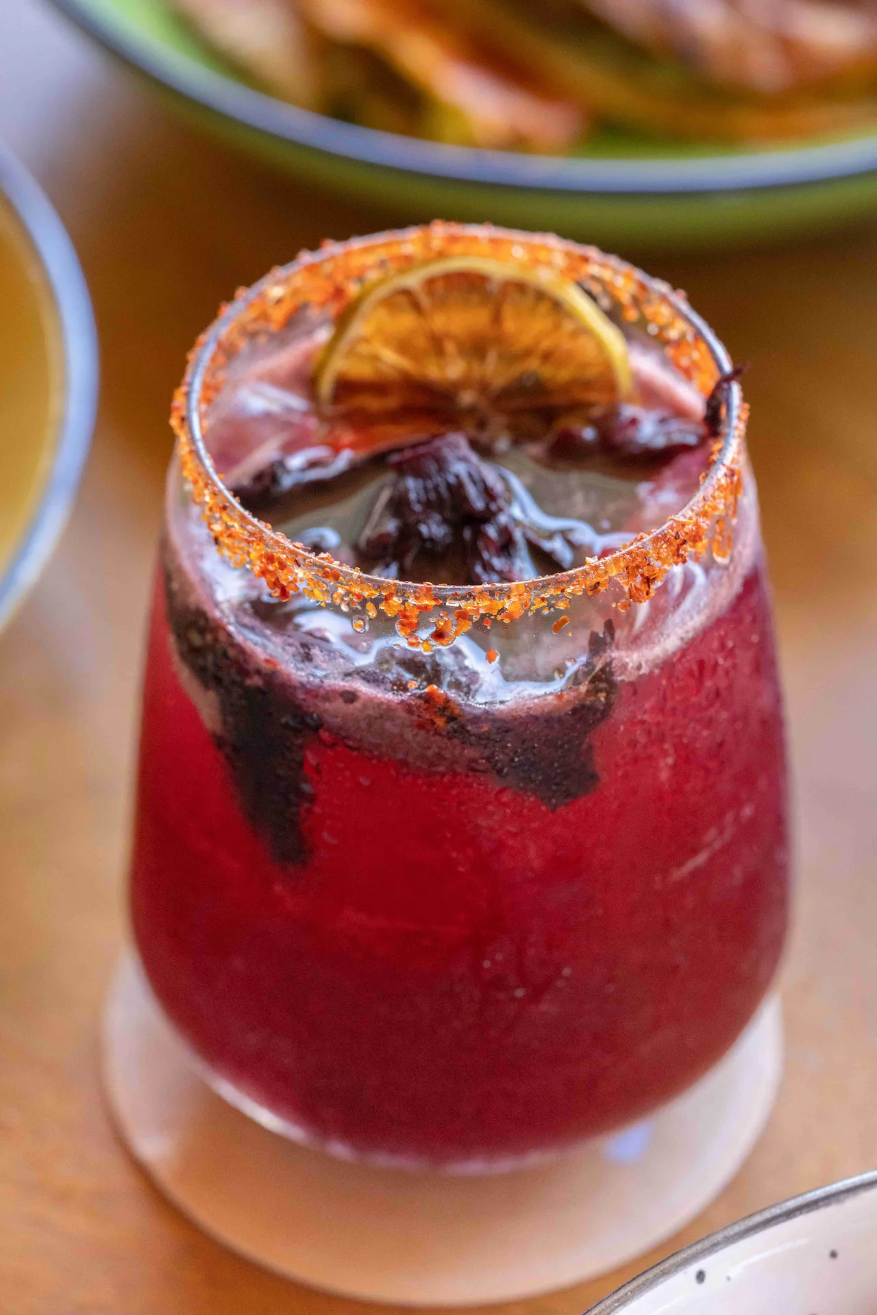 Iced hibiscus drink with chili rim, garnished with dried citrus and flowers in a tropical dining setting.
