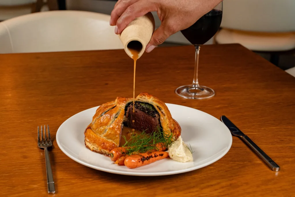 Beef Wellington plated with roasted carrots and mashed potatoes, as sauce is poured over the golden pastry.