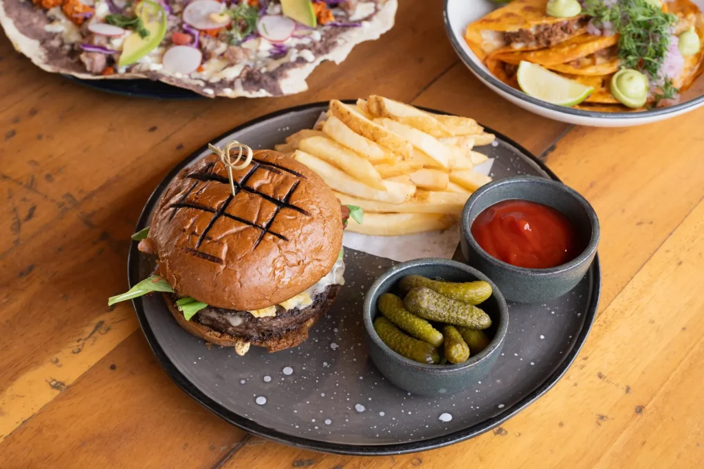 Grilled burger with fries, pickles, and ketchup, alongside tacos and tostadas with vibrant toppings.