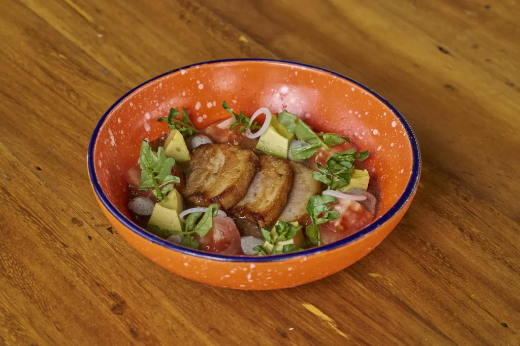 Pork belly slices with avocado, tomato, onion, and herbs in a savory broth, served in an orange bowl.