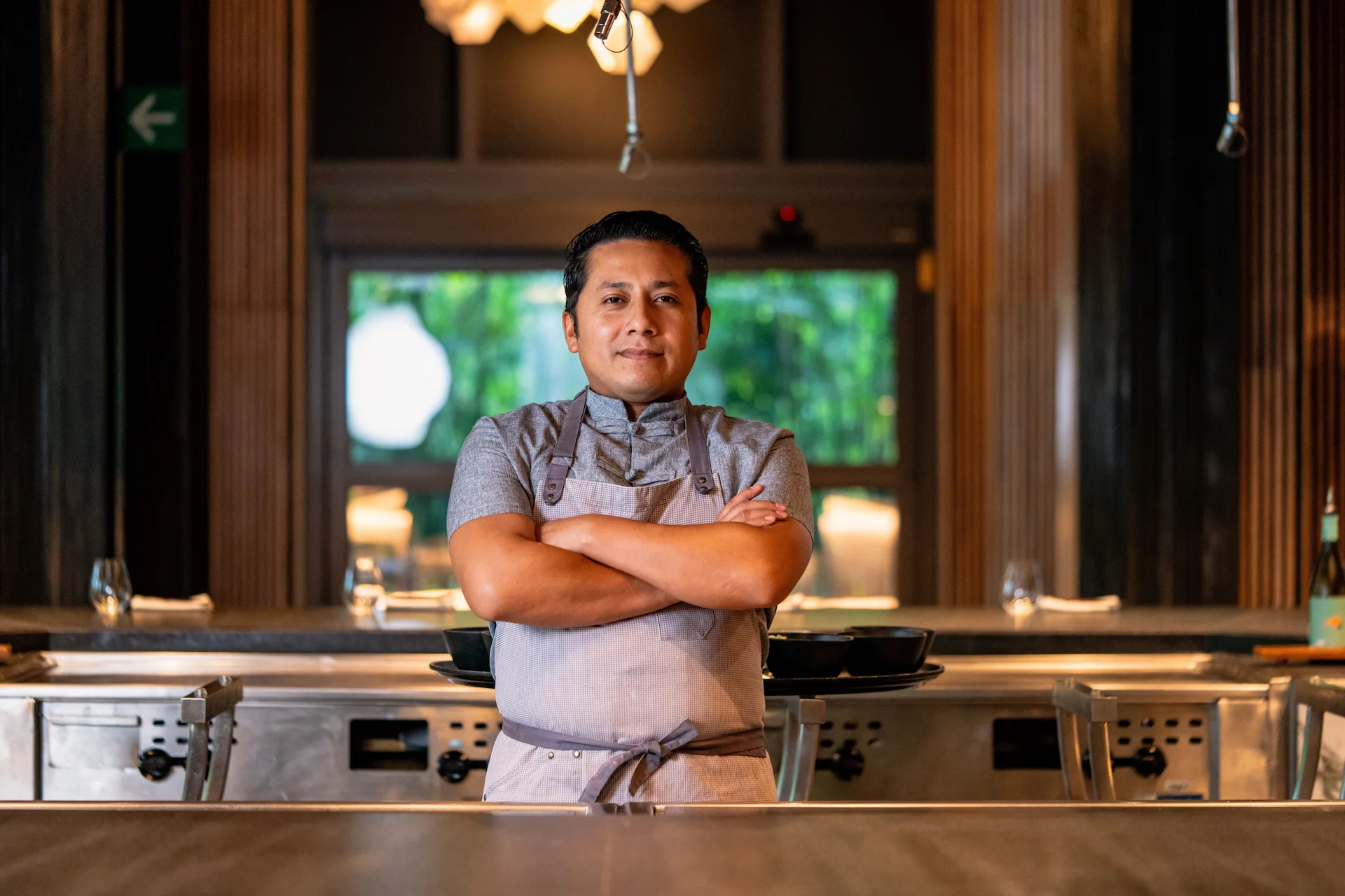 Chef Thare Osorio in grey uniform, arms crossed in Kengai’s kitchen with warm lighting and upscale decor.