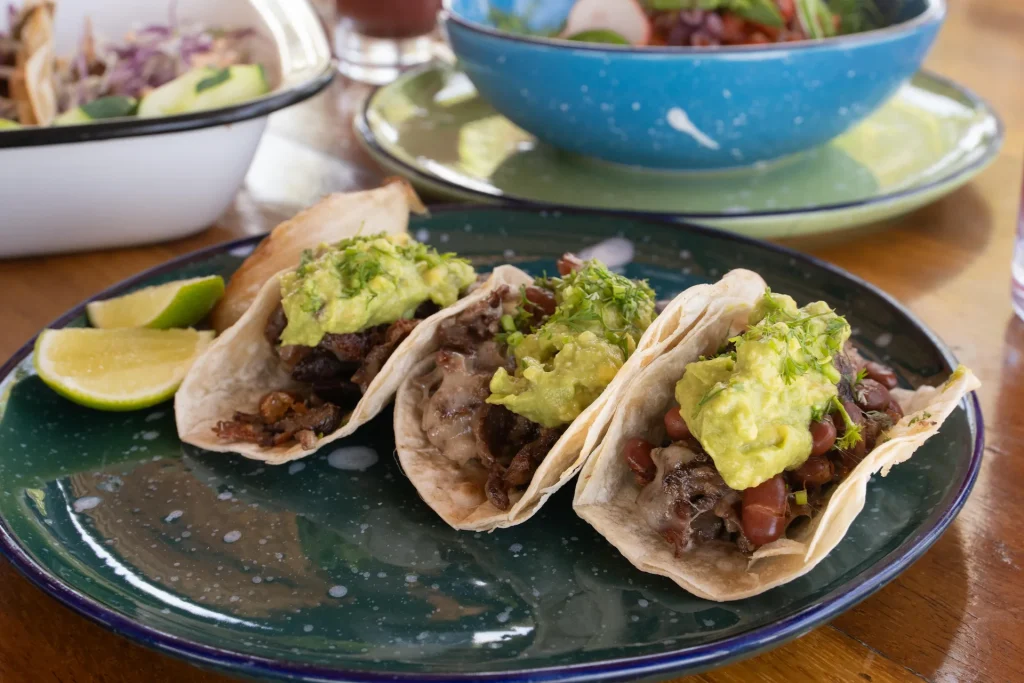 Three tacos with guacamole and herbs on green plate at CHIRINGUITOS, with lime wedges and shared dishes in background.