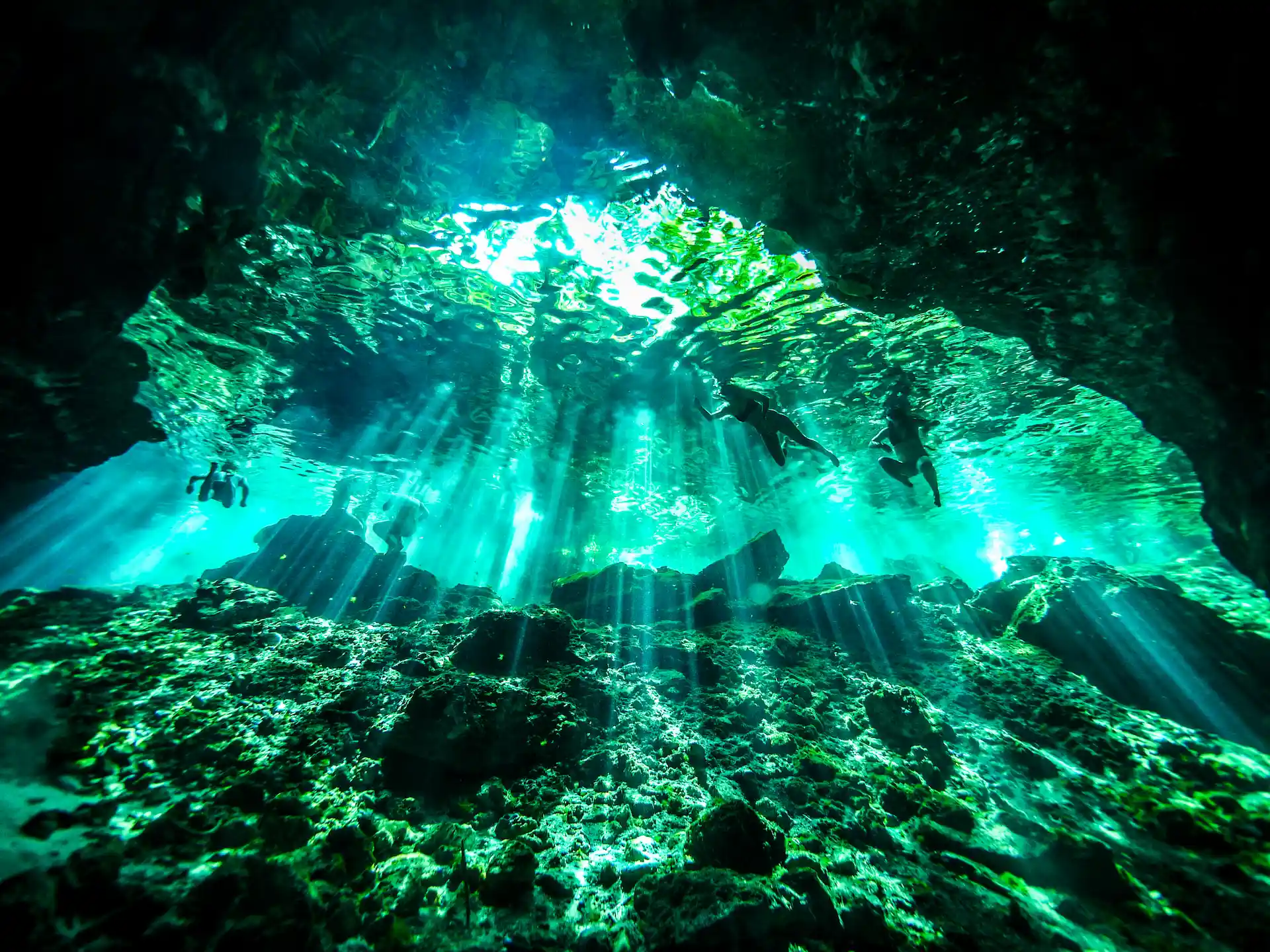 Snorkelers swimming in turquoise cenote with sunbeams illuminating rocky underwater cave floor
