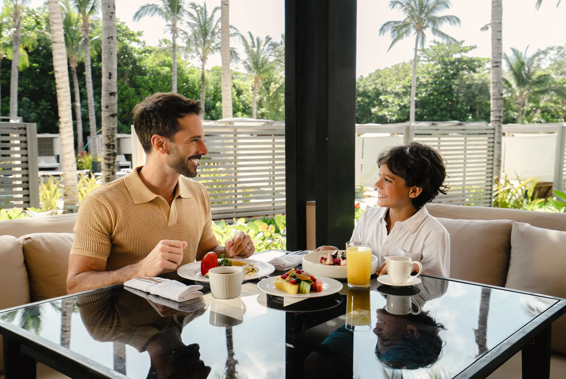 Two guests share breakfast outdoors at a tropical resort, surrounded by palm trees and morning light.