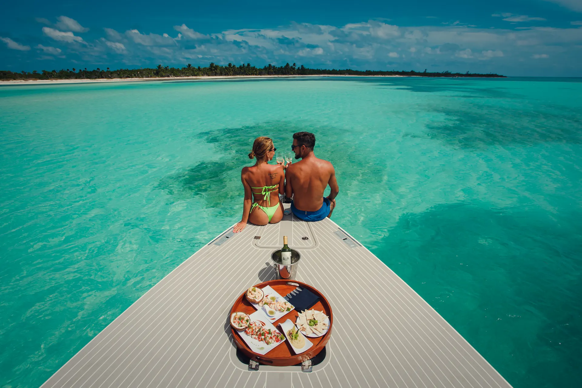 Couple seated on boat bow with wine and tropical lunch, overlooking turquoise water and palm-lined shore.