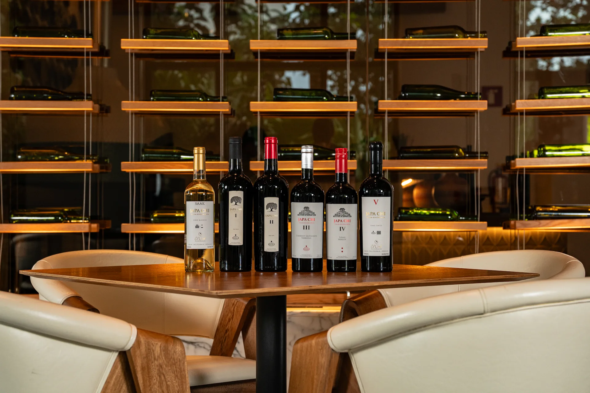 Elegant wine tasting setup featuring six JAPA CHI bottles on a wooden table, framed by a wall rack of wine bottles.
