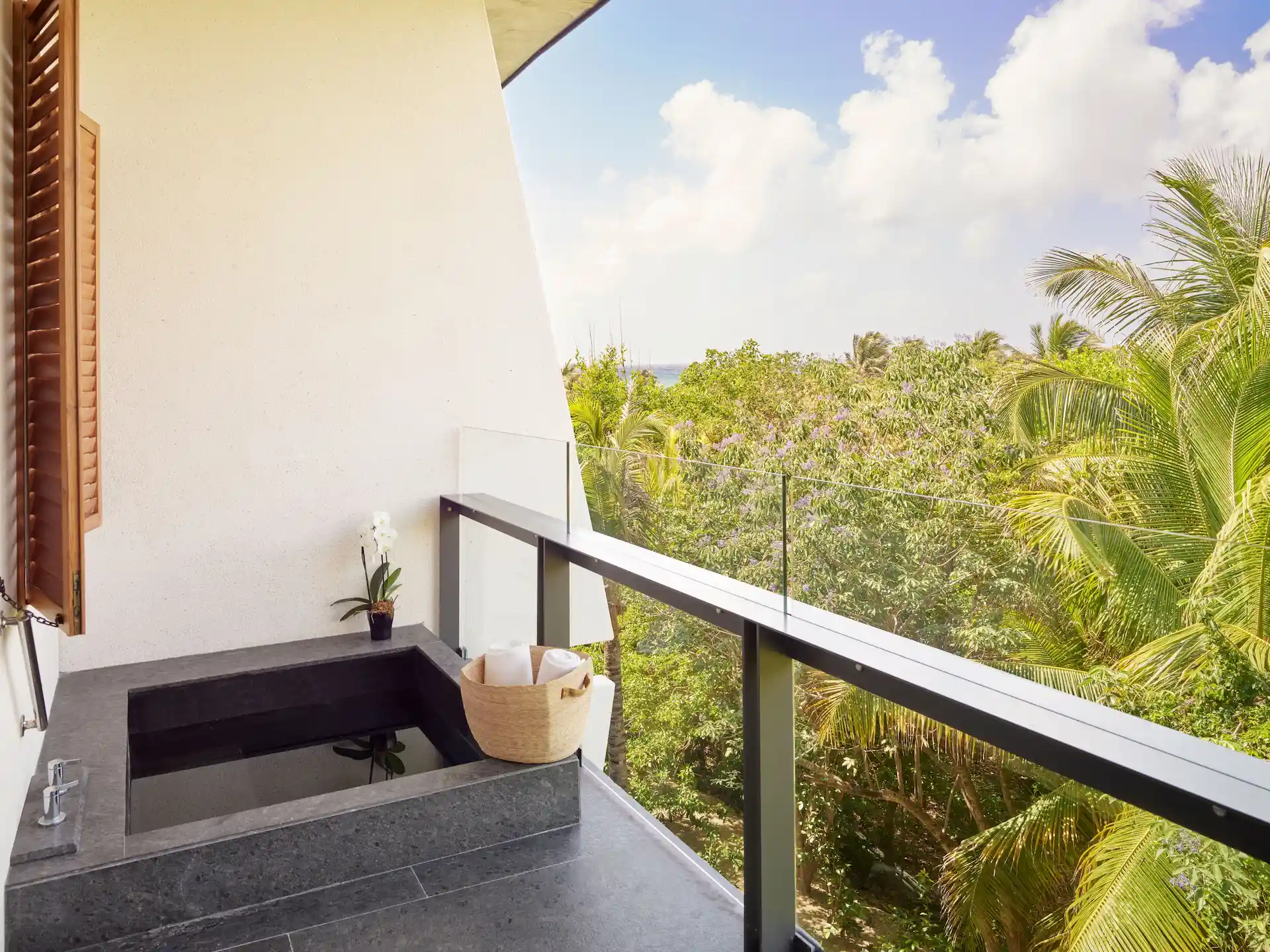 Balcony with dark stone soaking tub, orchid plant, and view of tropical greenery through glass railing