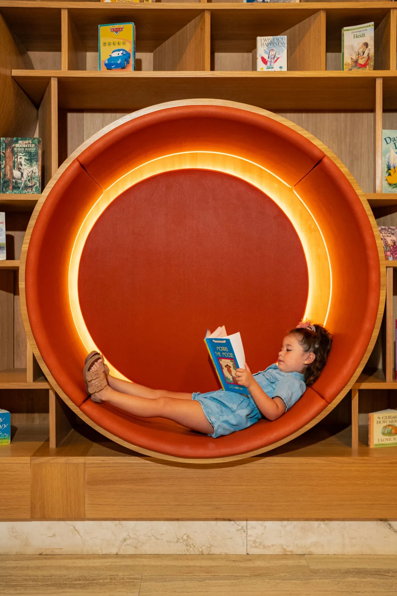 Child reads “Foss the Moose” inside a cozy, circular nook lined with orange cushions and soft light.