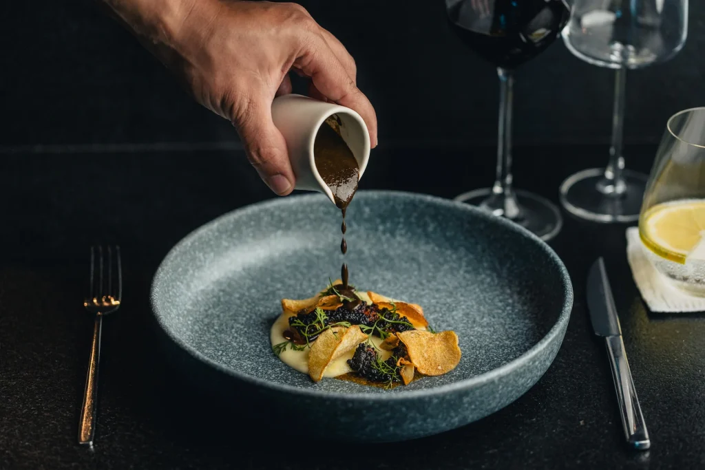 Gourmet dish at Conrad Tulum Riviera Maya with creamy base, crispy chips, herbs, and sauce being poured.