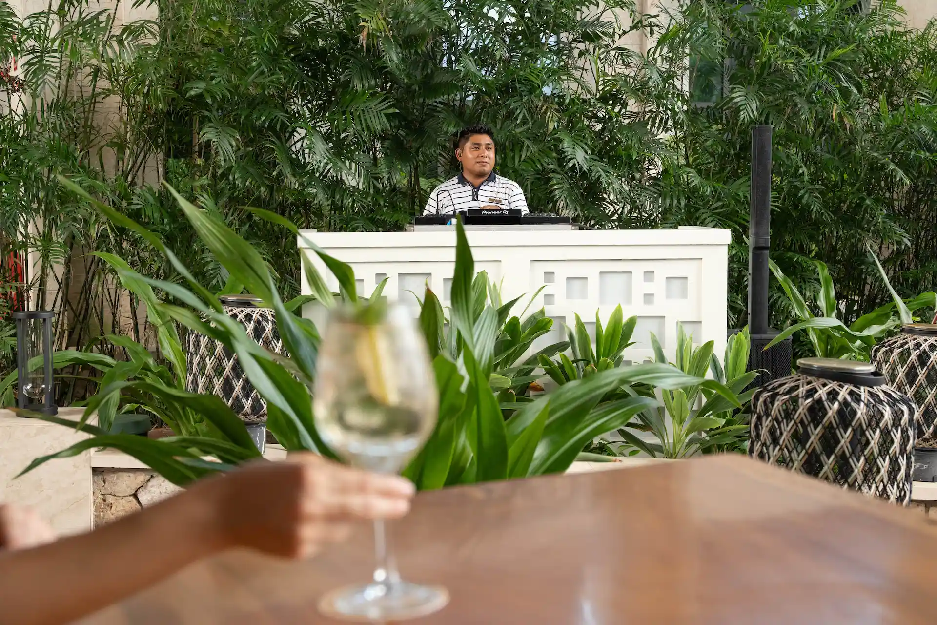 DJ behind white booth with tropical plants, wine glass in foreground, and woven lanterns in cozy setting