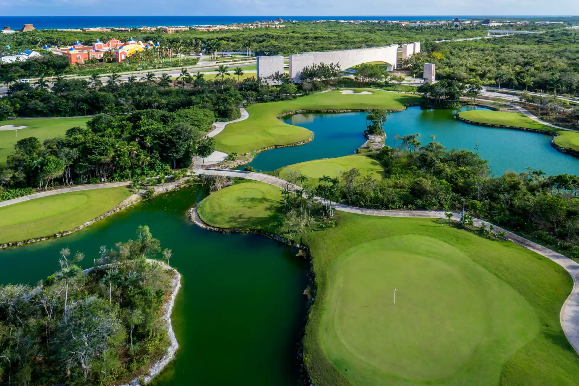Aerial view of tropical golf course with water features, resort buildings, and ocean in the distance