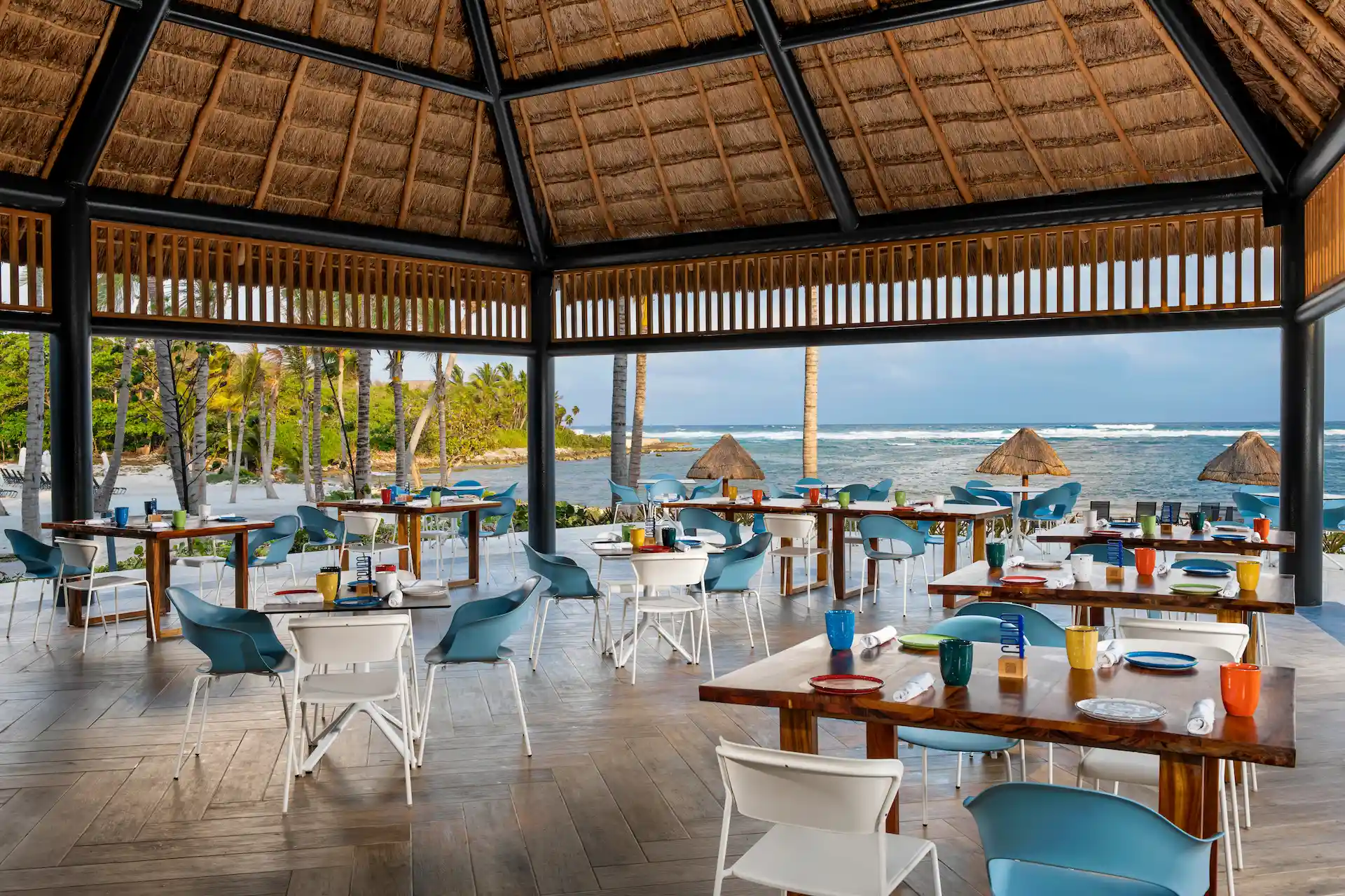 Beachfront restaurant with thatched roof, colorful table settings, and ocean view with palm trees