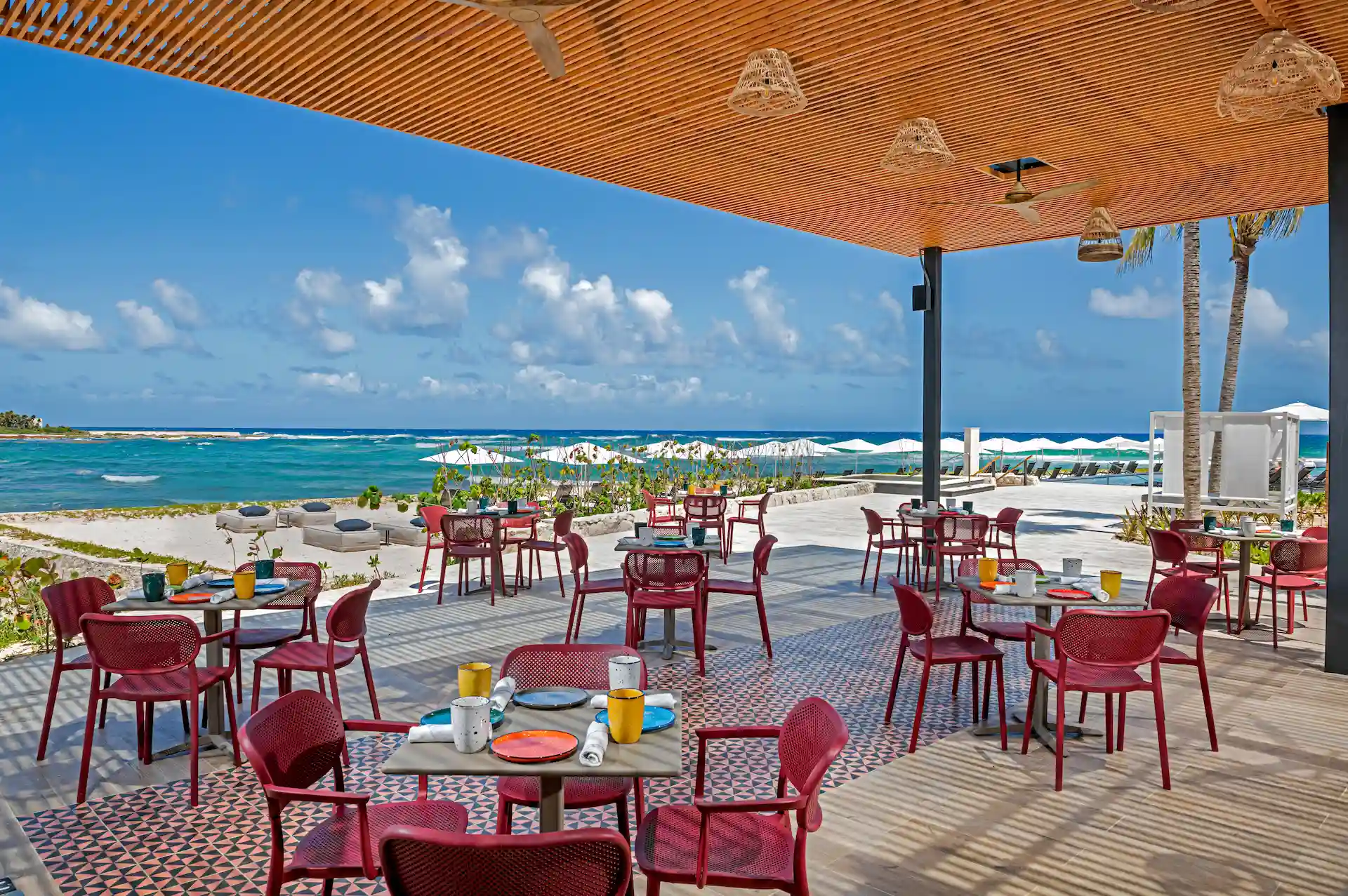 Beachfront restaurant with red chairs, wicker lights, and ocean view with lounge chairs and umbrellas