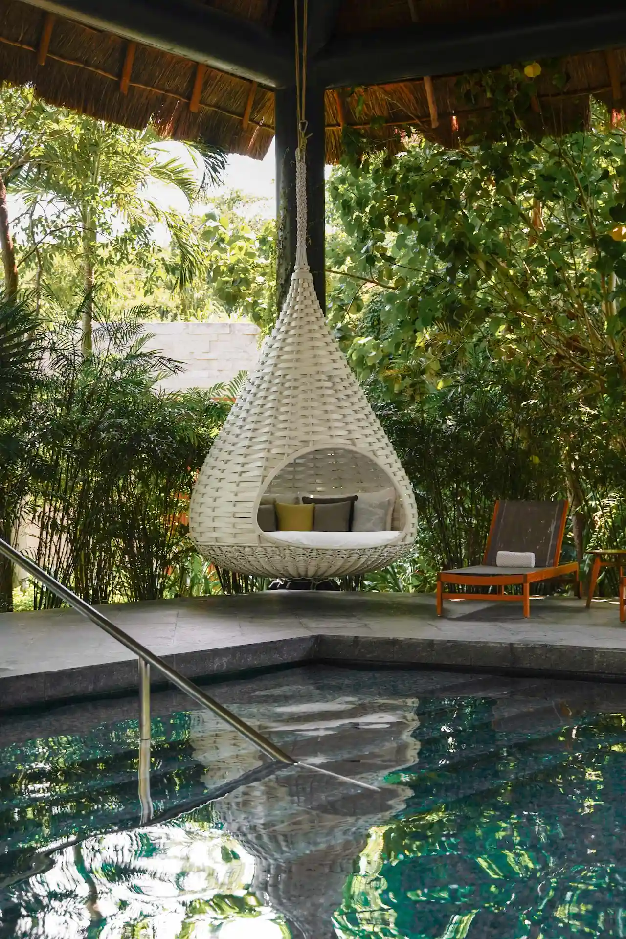 Hanging pod chair near pool with tropical plants, orange lounge chair, and thatched-roof structure