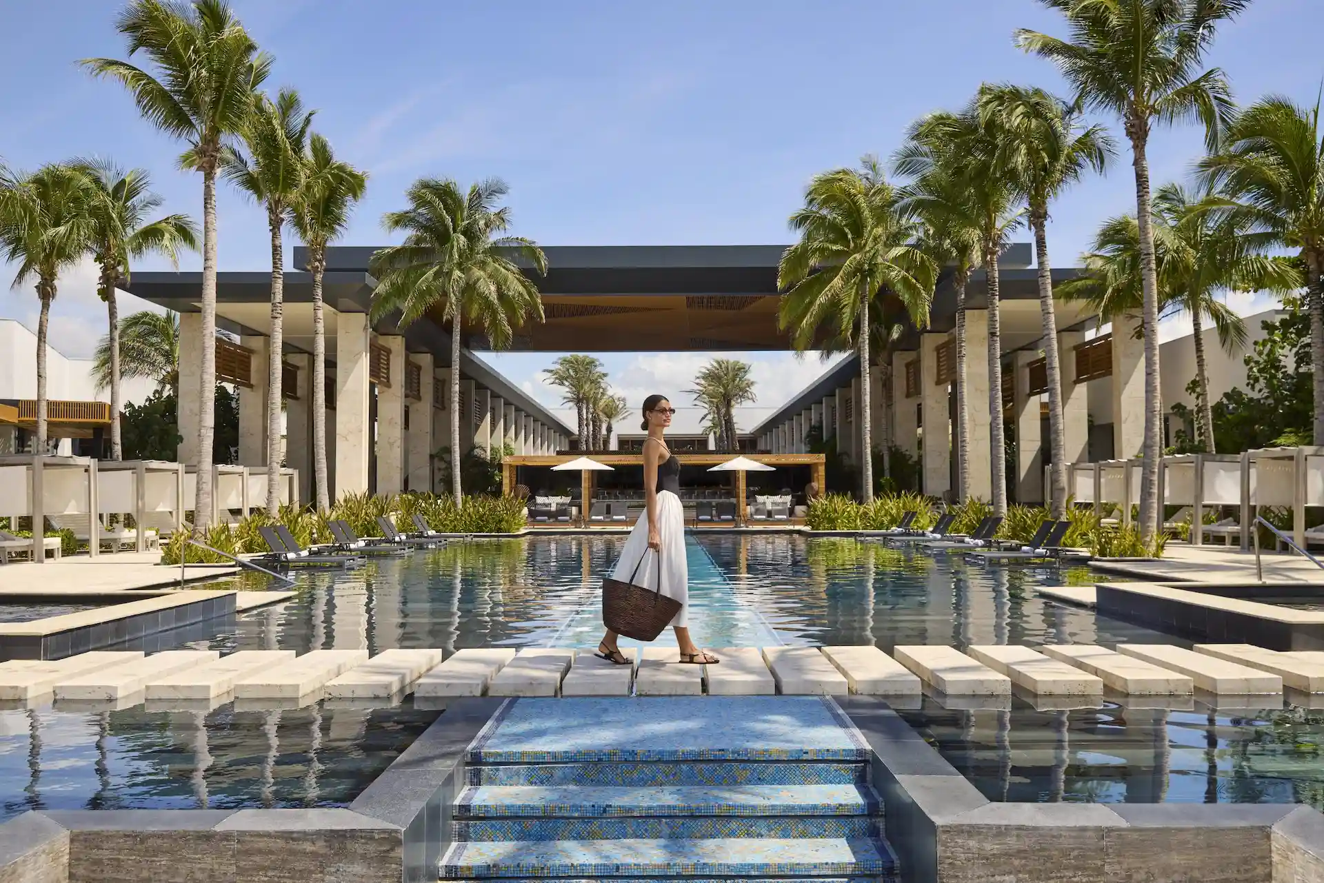 Woman walking across stone path over resort pool with palm trees, lounge chairs, and modern buildings