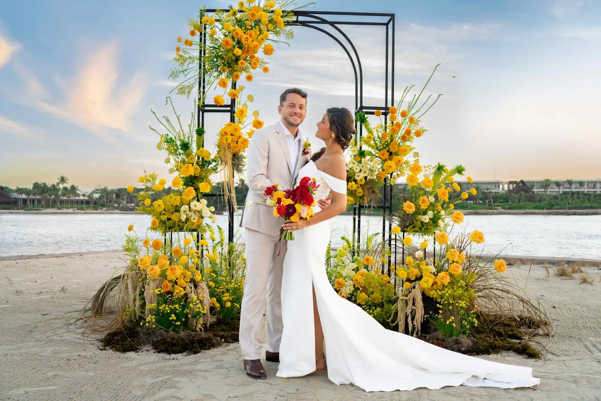 Couple standing under a vibrant floral arch during a beach wedding at sunset in Riviera Maya.