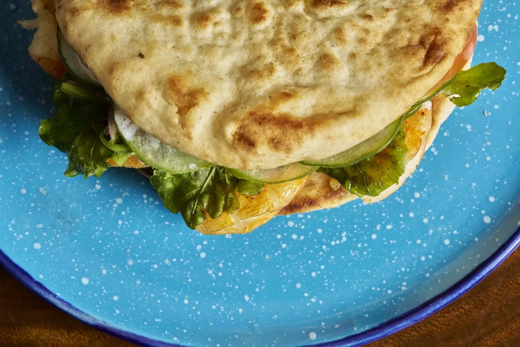 Grilled chicken flatbread sandwich with fresh greens, cucumber, and tomato on a speckled blue plate.