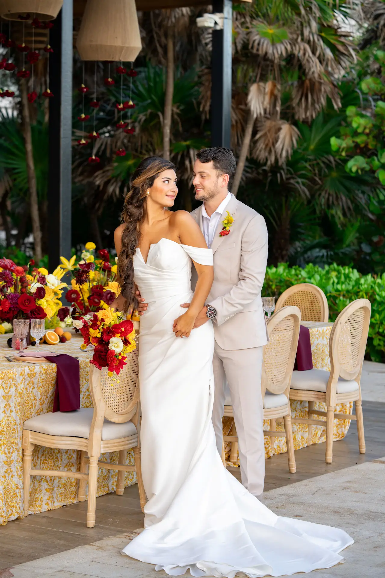 Bride and groom embrace beside a floral reception table, surrounded by lush greenery and festive decor.