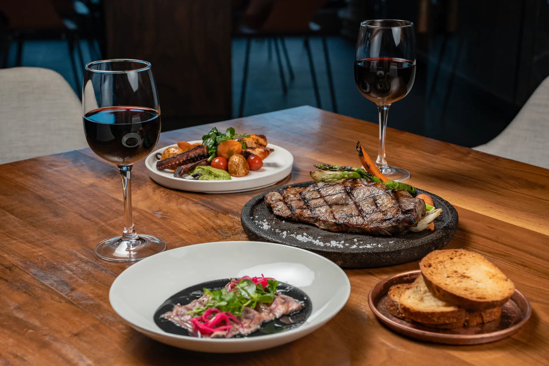 Gourmet spread with grilled steak, roasted vegetables, sliced meat with herbs, and red wine on a wooden table.
