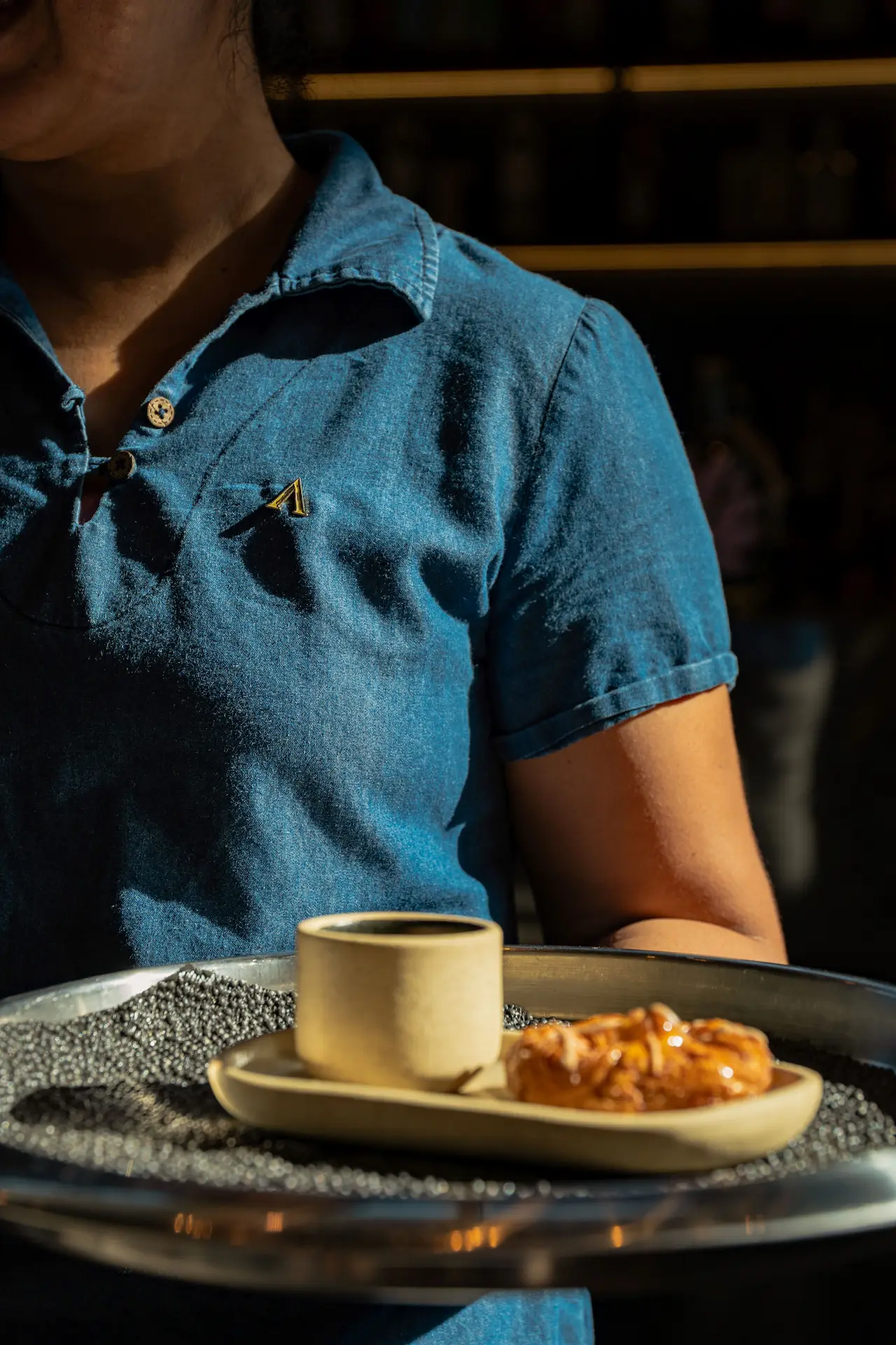 Server in denim shirt presents tray with coffee and nut-topped pastry on ceramic ware.