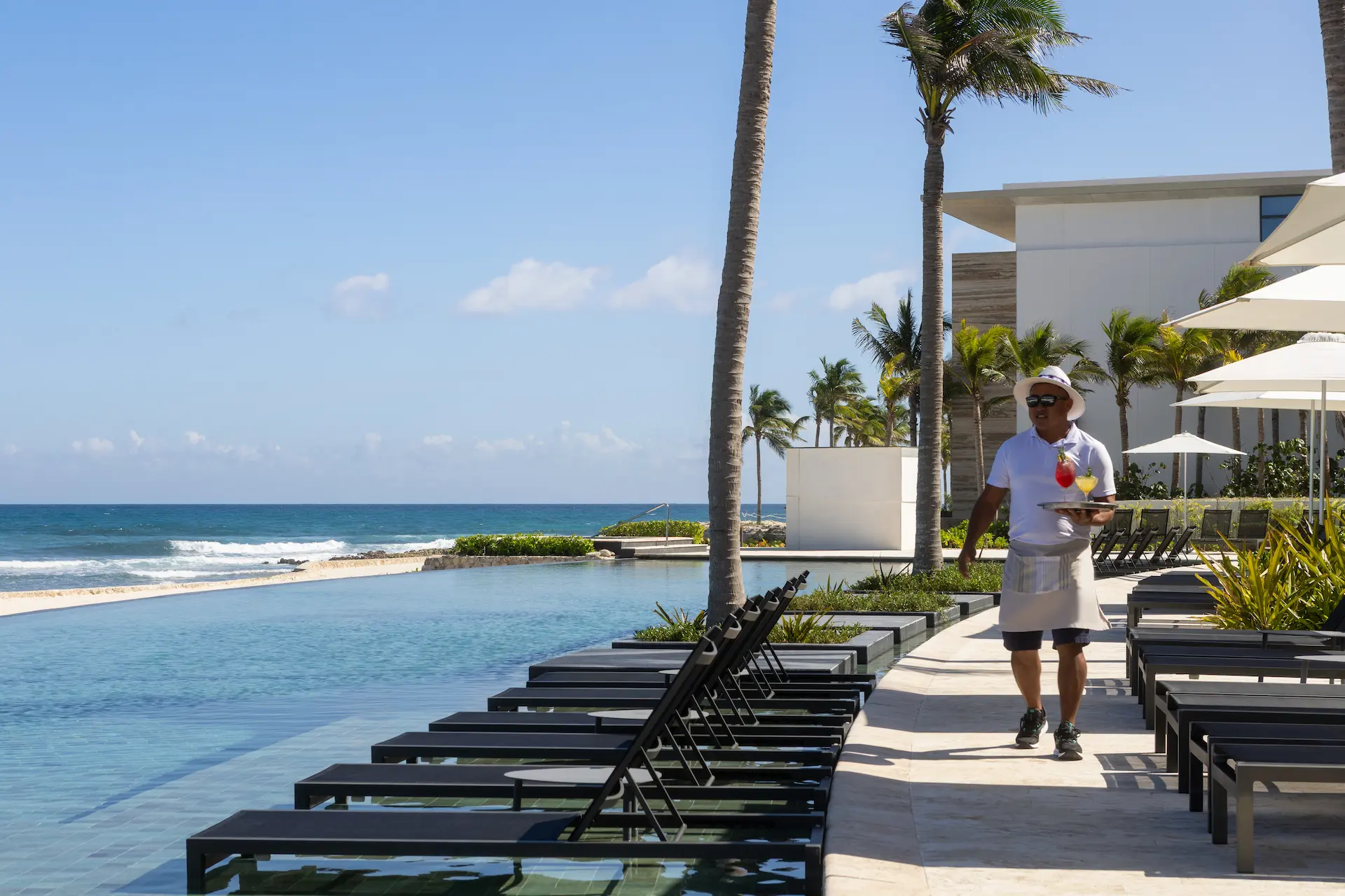 Chiringuito Leña waiter serves drinks by the infinity pool at beachfront resort. Palm trees and ocean in the background.