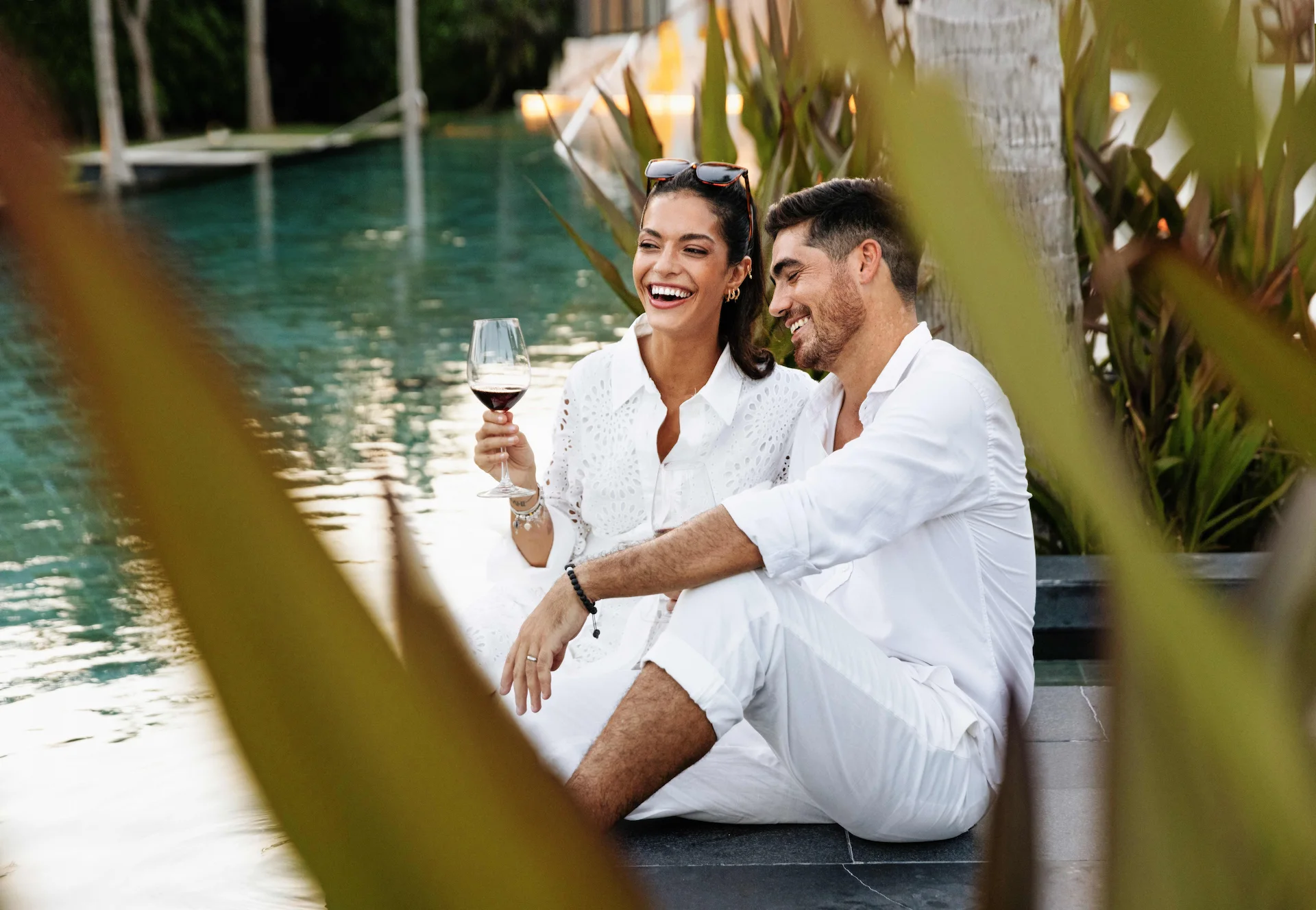 Two people in white relax by the pool, surrounded by tropical plants. One holds a glass of red wine.