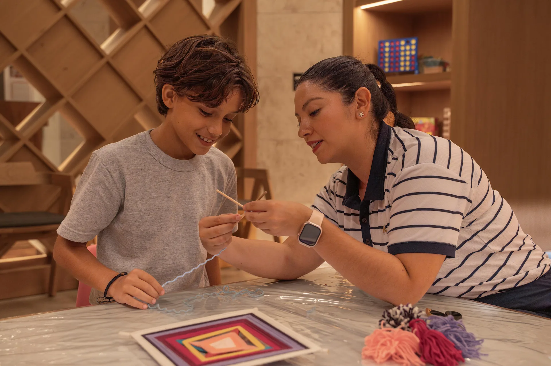 Woman and child create yarn art together at a craft table, surrounded by colorful threads and geometric designs.