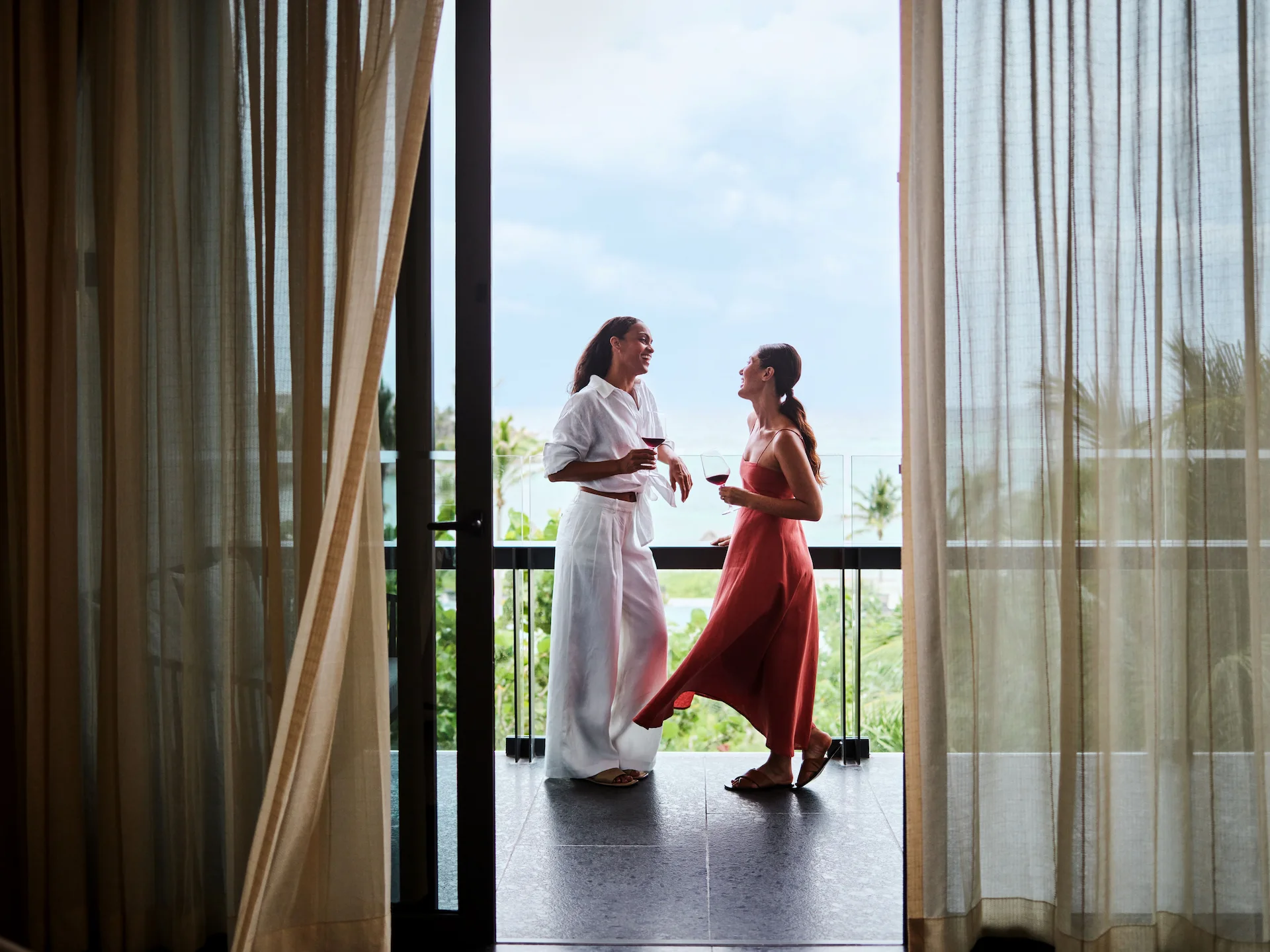 Two people enjoy drinks on a resort balcony, one in red dress, overlooking tropical landscape and ocean.