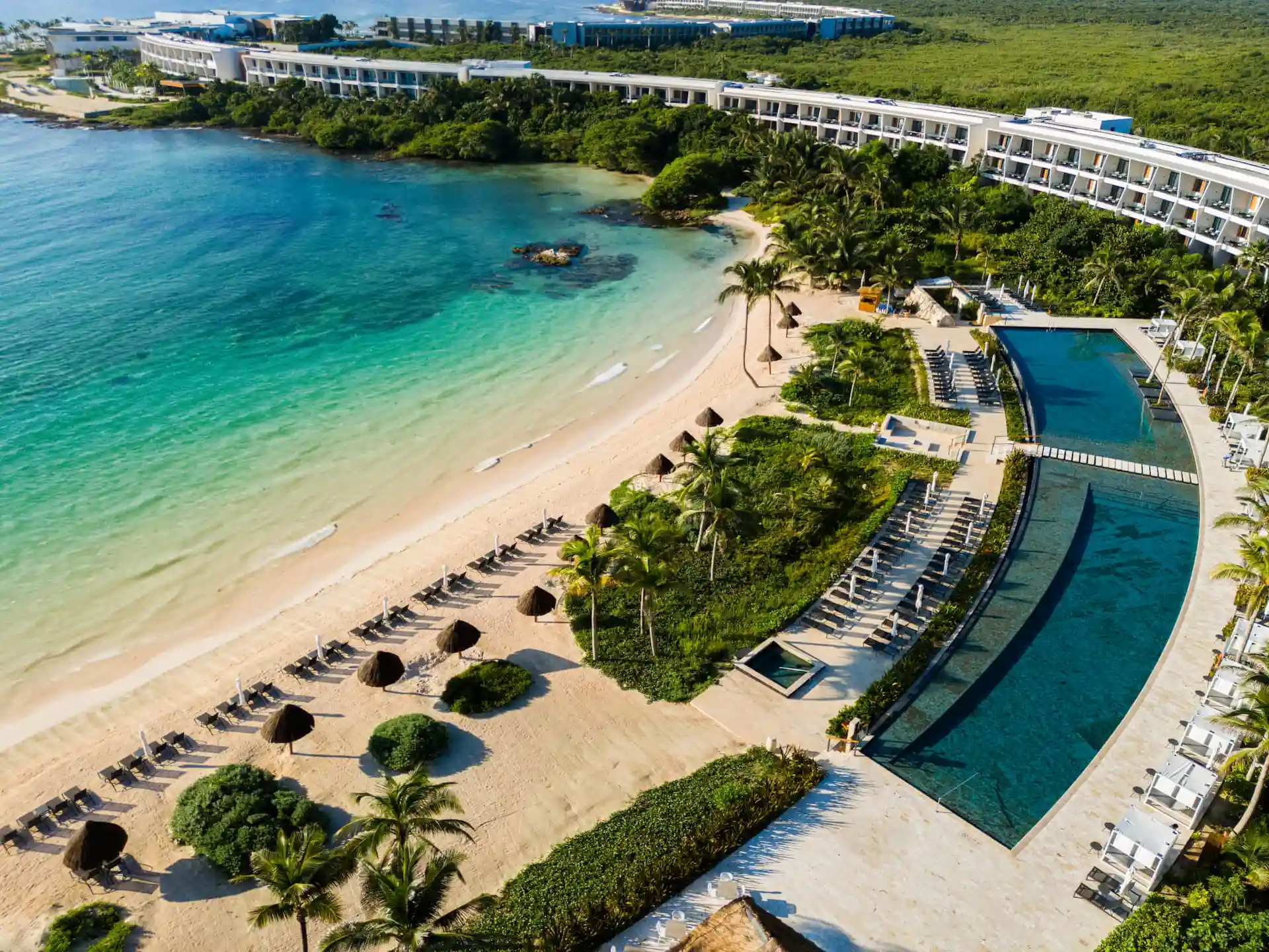 Vista aérea de un complejo turístico frente al mar con edificios, palmeras, una piscina infinita y hileras de tumbonas en una playa de arena blanca.