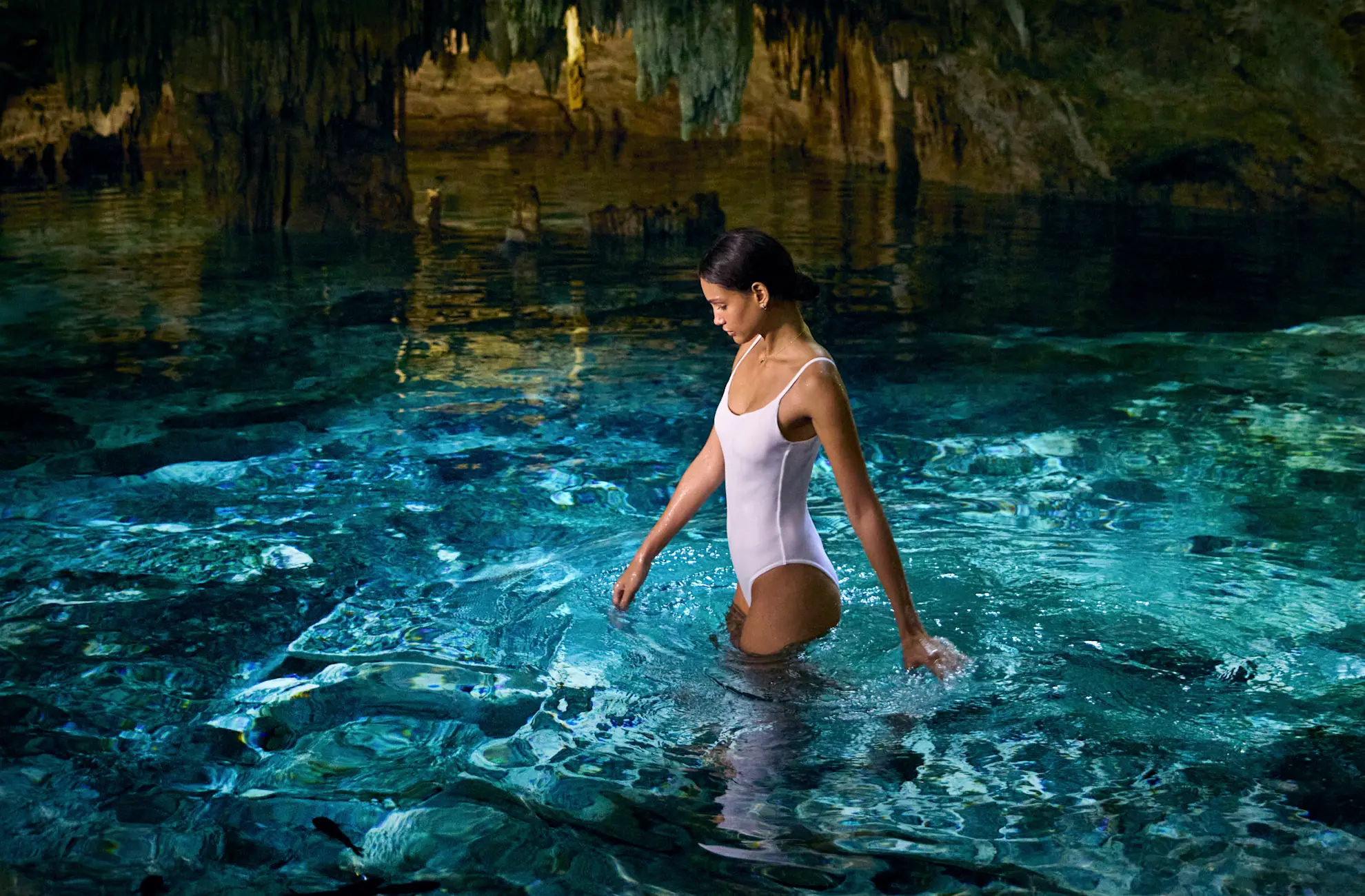 Mujer parada en una piscina subterránea, con luz entrando por una grieta en la roca de arriba